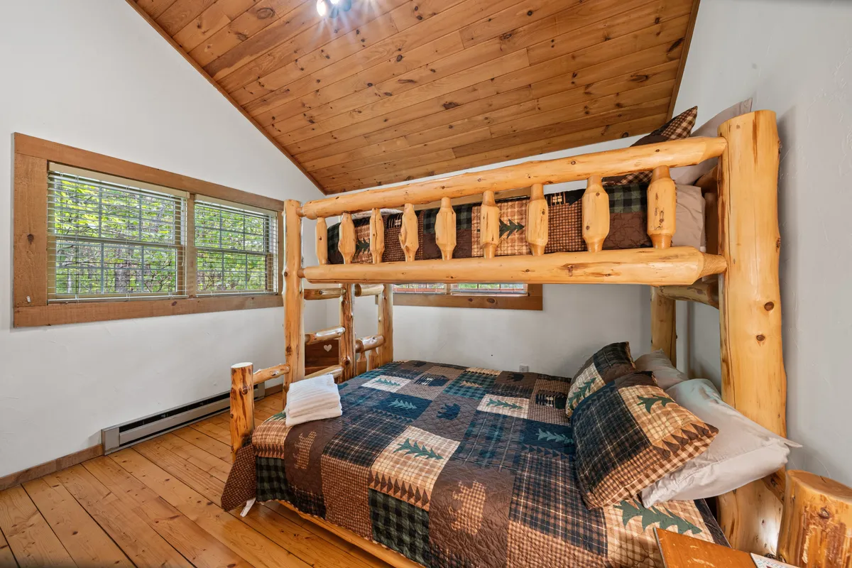 A charming bunk room with a solid wood bunk bed topped with a colorful patchwork quilt, wooden beams overhead, and windows revealing greenery outside. The space is bright and playful, perfect for children or multi-generational guests.
