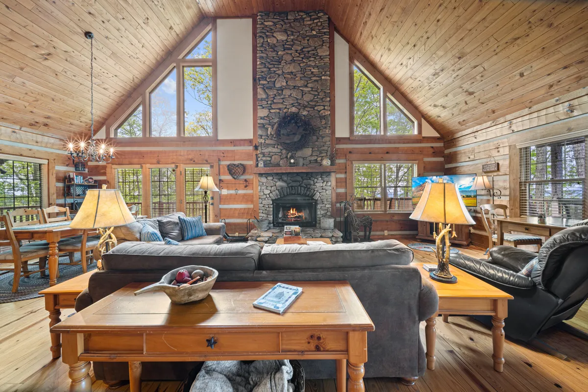 A warm and welcoming great room featuring a grand stone fireplace, tall wooden beams stretching to a vaulted ceiling, expansive windows letting in abundant natural light, and comfortable seating arranged for socializing and relaxation. The view through the windows reveals glimpses of the surrounding mountain landscape, while the spacious layout creates a sense of openness and togetherness.