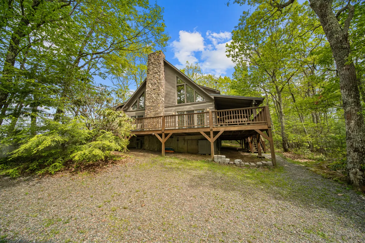 This image features a wooden deck with a stone chimney and is surrounded by lush trees, creating an inviting transition between the home and the natural outdoors. The wide deck space suggests room for pets and people alike to gather, relax, and enjoy the peaceful forested setting.