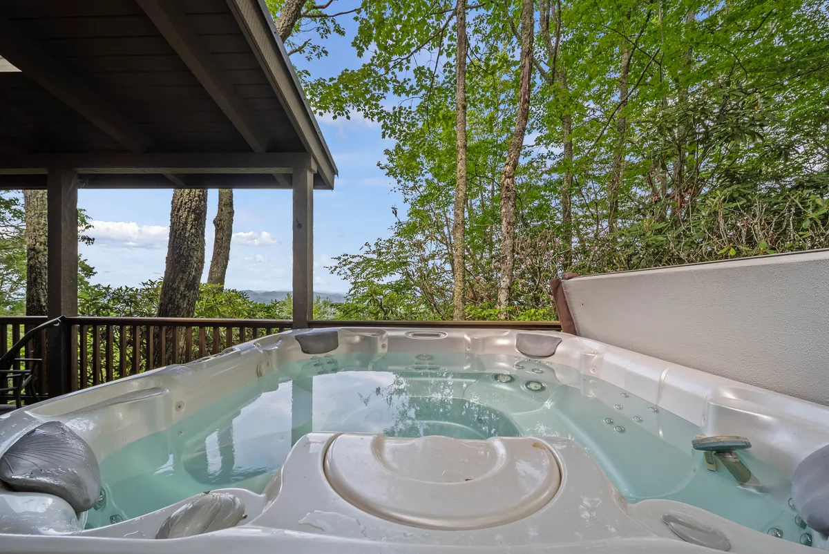 This exterior image features a spacious hot tub set against a backdrop of lush trees and open sky. The serene setting highlights the tranquil natural environment surrounding the cabin, with the hot tub as a central, inviting element. The view extends into the woods, emphasizing privacy and immersion in nature—ideal for post-adventure relaxation.