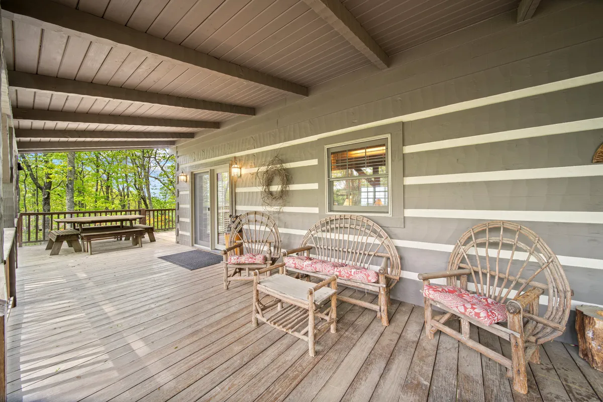 A welcoming wooden deck is furnished with comfortable wicker chairs and an outdoor dining table, set under open skies. The deck looks out over the landscape, suggesting a perfect spot for relaxing, socializing, or simply appreciating the mountain air and sweeping views. Outdoor elements like railings and distant treetops connect the living space seamlessly to nature.